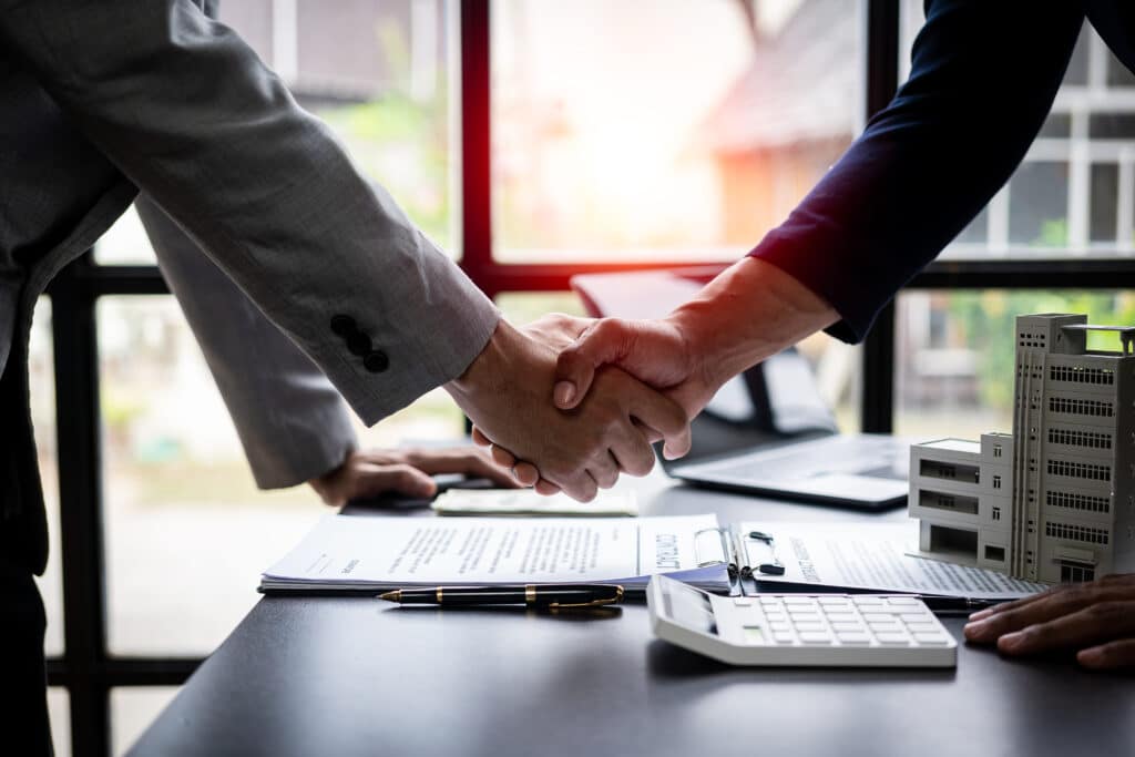 Poignée de main sur contrat : signature accord immobilier Poignée de main scellant un contrat immobilier, avec un stylo, une calculatrice et une maquette de bâtiment sur un bureau.