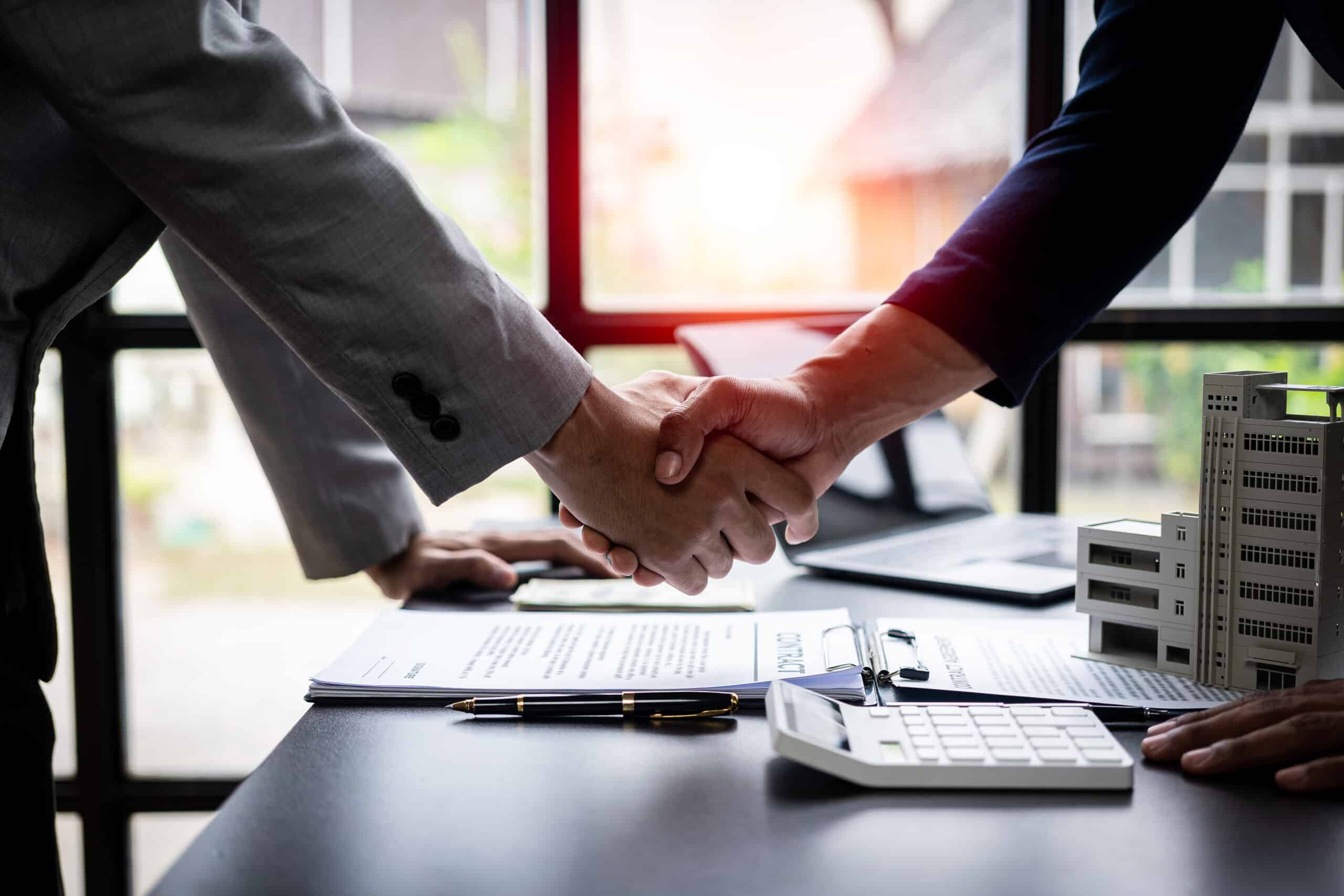 Poignée de main scellant un contrat immobilier, avec un stylo, une calculatrice et une maquette de bâtiment sur un bureau.