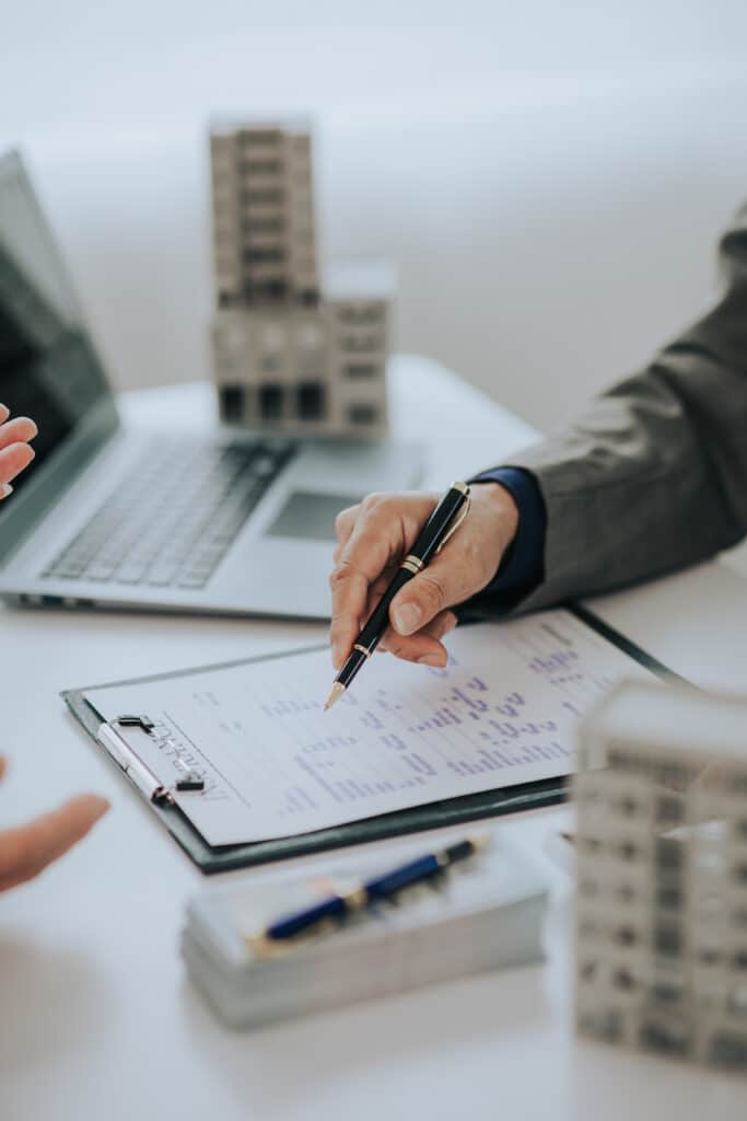 Signature d'assurance : transaction immobilière en bureau Main tenant un stylo au-dessus d'un document "INSURANCE" sur un presse-papiers, avec des maquettes de bâtiments et un ordinateur portable.