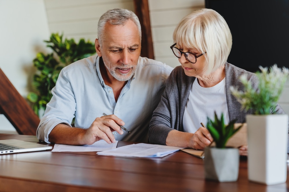 Couple senior : Planification financière et gestion de retraite Couple âgé planifiant ses finances ou sa retraite. L'homme explique les documents à la femme qui écrit à la table en bois.